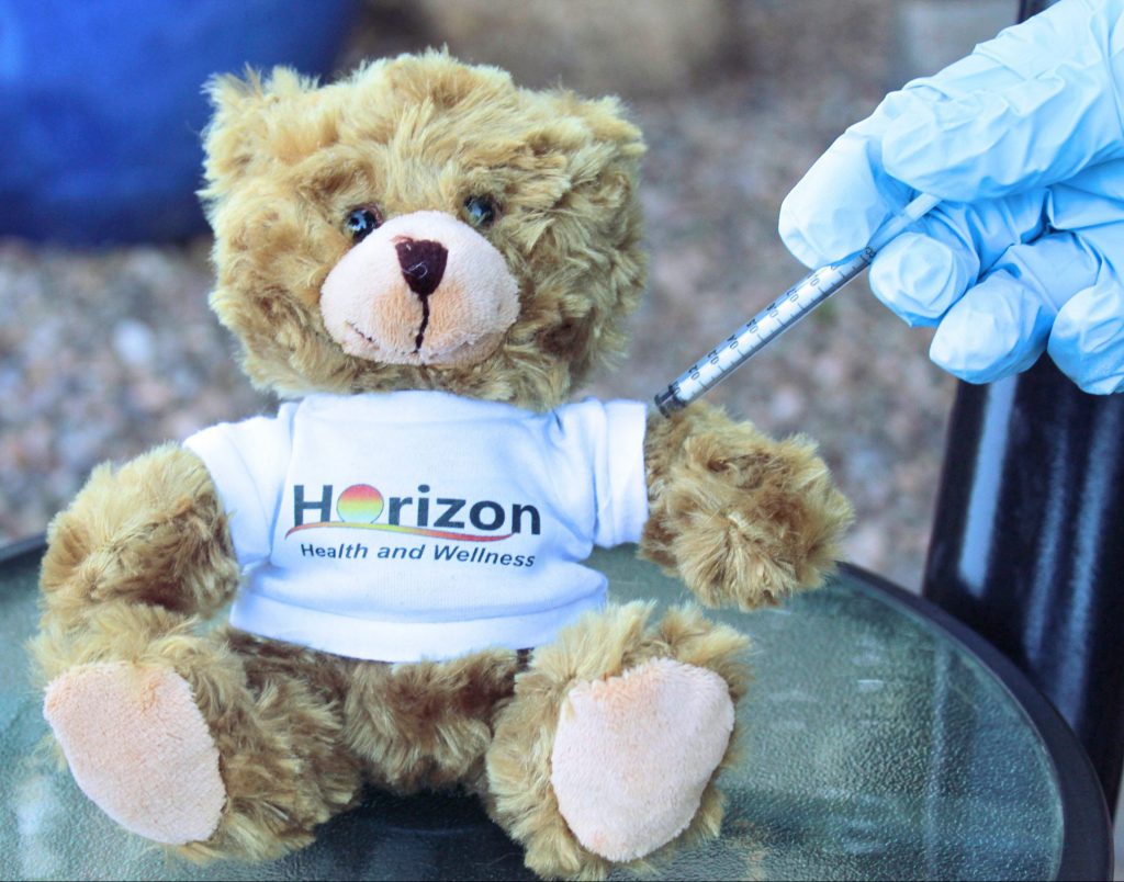 A hand with a glove giving a brown, stuffed bear wearing a Horizon Health and Wellness shirt the COVID vaccine in its arm.