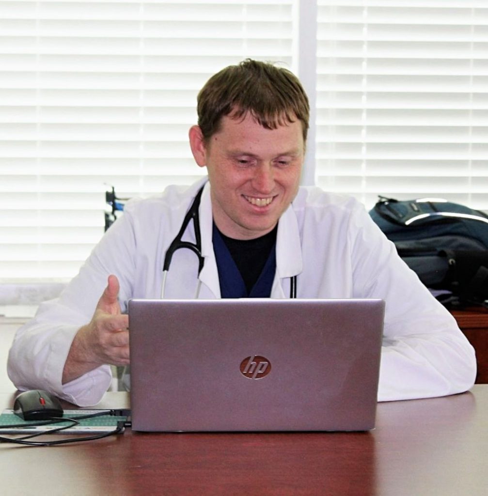 A smiling Physician Assistant with a stethoscope having a Telemedicine appointment on his laptop at his desk.