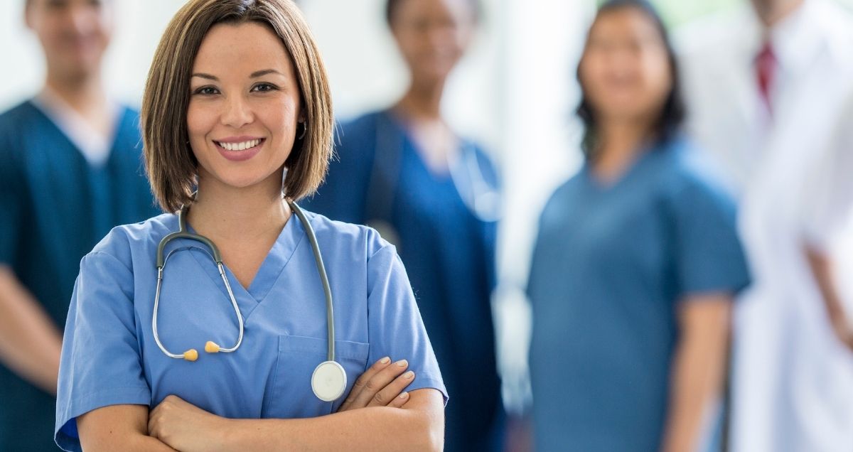 A Hispanic doctor in scrubs with her hands crossed standing confidently in front of a group of healthcare workers.