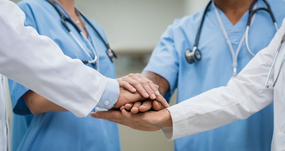 Four cooperative doctors in scrubs and lab coats huddled together in a circle with their hands together.