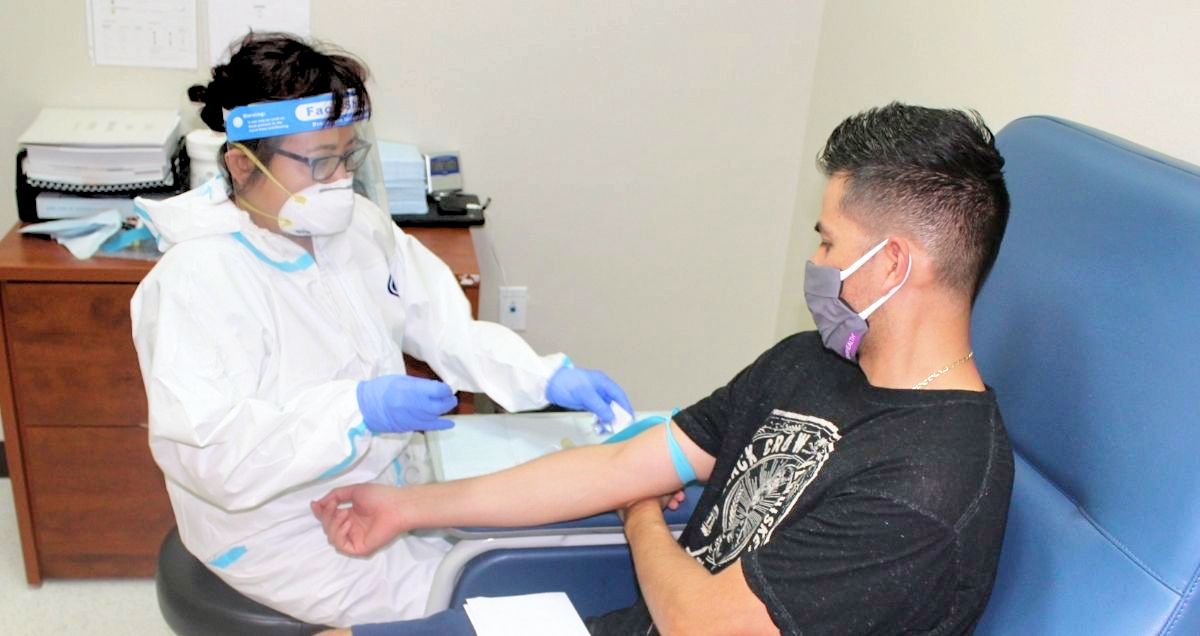 A nurse wearing a face shield and glasses giving the COVID vaccine to a male patient in an office.