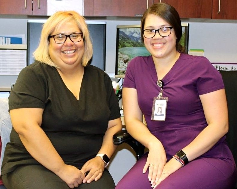Two smiling Hispanic nurses in scrubs sitting at their desks surrounded by computers in the office.