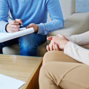 A therapist writes on a notepad as he talks to his patient sitting with clasped hands.
